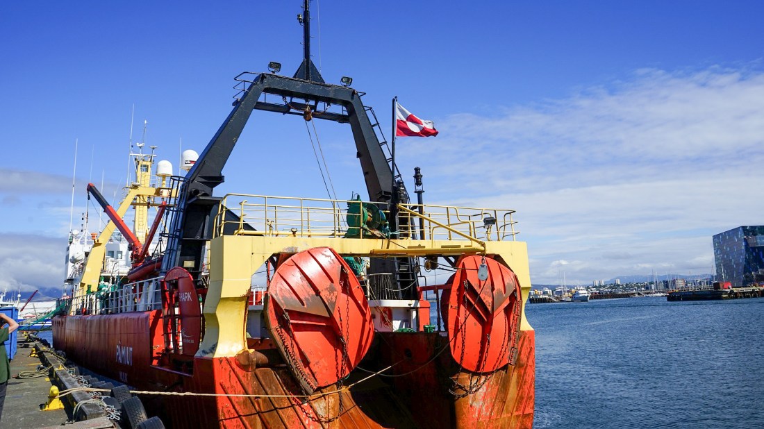 Commercial whaling vessel docked at a harbor, showing large red circular harpoon reels and a yellow A-frame structure mounted on deck. The ship flies a Greenlandic flag and has industrial whaling equipment visible. The harbor scene includes a modern city skyline with a distinctive building visible in the background across calm waters on a partly cloudy day. Image by Bernd Hildebrandt from Pixabay.