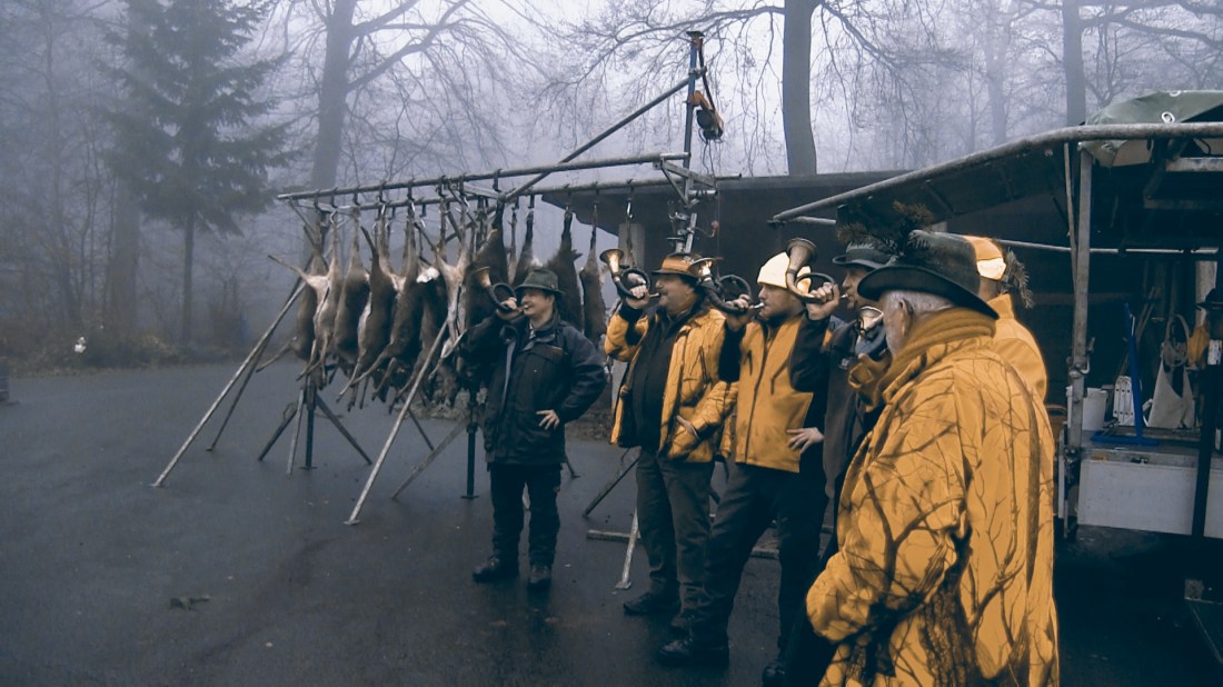 A group of hunters standing in a line wearing yellow hunting jackets and dark trousers, with some wearing hunting hats. Behind them hangs a row of harvested rabbits or similar game animals on a wooden frame. The scene is set in a misty or foggy wooded area, creating a moody atmosphere. The hunters appear to be gathered for what might be an end-of-hunt gathering or inspection of their quarry.