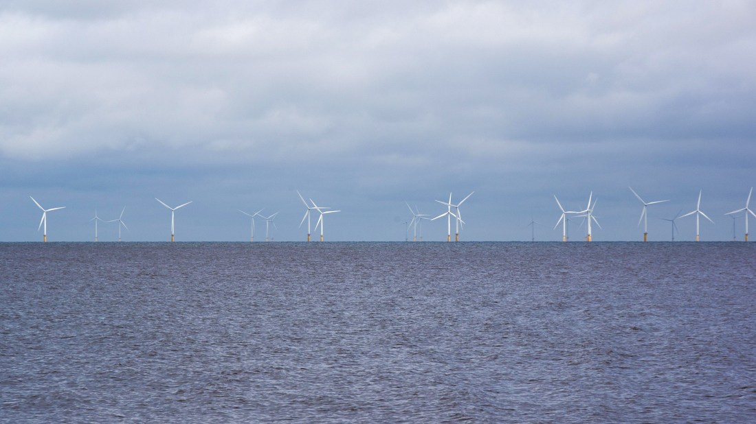 The image shows an offshore wind farm with multiple white wind turbines arranged in a line across the horizon. The turbines are installed in the sea, with their bases partially submerged in the grey-blue water. The sky above is overcast with a light grey cloud cover, creating a moody atmosphere. Each turbine has three blades and appears to be of significant size, standing tall above the water's surface. The turbines are evenly spaced and extend across the entire width of the frame, demonstrating the scale of the offshore wind energy installation. Image by Julia Schwab from Pixabay