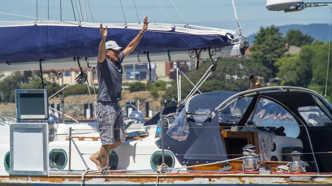 The image shows a person standing on a sailing yacht with their arms raised upwards in what appears to be a joyful or celebratory gesture. They're wearing a light-coloured cap, a grey t-shirt and patterned shorts. The yacht has a dark blue sail cover folded on the boom above. The vessel has a white hull with porthole windows and various sailing equipment visible, including winches and rigging. There appears to be another person partially visible inside the cabin area. The scene takes place on a sunny day with a shoreline and buildings visible in the background, suggesting this is at a marina or coastal area.