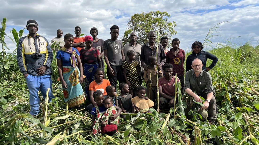 A group photo of approximately 20 Black people from a rural African community, including adults and children, standing and sitting together in a lush agricultural field. The group appears to be a farming community, with people of various ages wearing casual clothing in bright colours including orange, blue, yellow, and traditional patterned fabrics. In the foreground and surrounding the group are green crops including what appears to be maize plants and other vegetables. The setting suggests a rural farming area with rich, fertile soil. On the right side of the group stands a white journalist with glasses wearing olive-green clothing. The sky overhead is partly cloudy, and there are trees visible in the background. The image captures what appears to be a community gathering or visit to showcase local agricultural work.