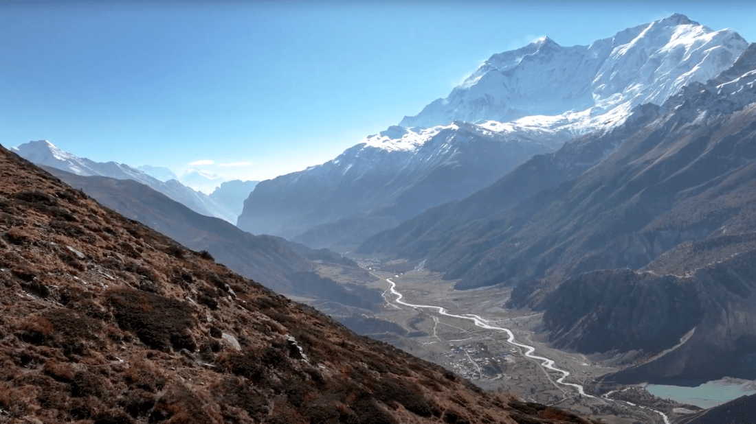 A dramatic mountain valley landscape showing snow-capped Himalayan peaks in the background with a glacial river winding through a broad valley floor below, surrounded by steep, barren mountainsides covered in sparse vegetation.