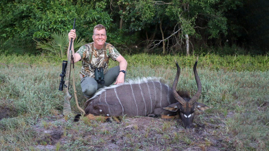 A hunter in camouflage clothing and glasses kneels beside a harvested nyala antelope in a grassy field. The hunter holds a rifle and poses with the animal, which displays distinctive white vertical stripes on its dark brown coat and long spiral horns. Dense vegetation is visible in the background.
