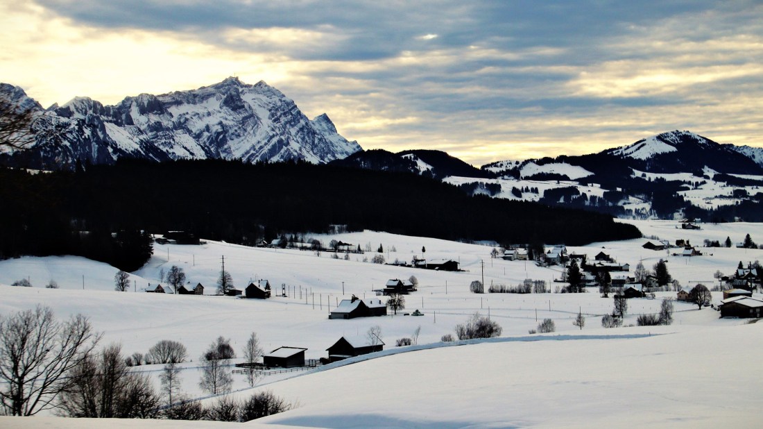 Winter landscape showing a snow-covered alpine valley with traditional wooden chalets scattered across rolling hills. Dramatic snow-capped mountain peaks rise in the background beneath a cloudy sky with soft golden light. Bare trees dot the foreground of the pristine white valley.