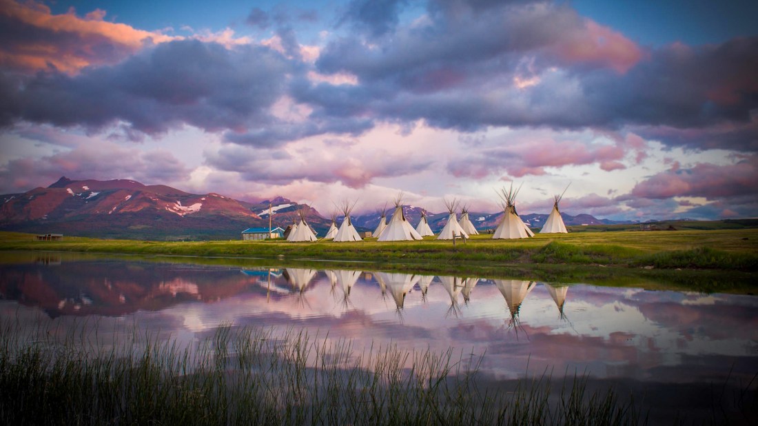 A row of traditional Native American tipis with white canvas covers stands on a grassy plain at sunset, their pointed wooden poles extending skyward. The tipis are perfectly reflected in the still waters of a lake in the foreground, where reeds grow along the shore. Behind them, snow-capped mountains rise against a dramatic sky filled with pink, purple, and grey clouds illuminated by the setting sun. A small blue building sits to the left of the tipis on the open prairie.